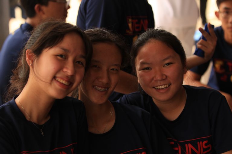Three ACS(I) tennis team members in Tennis 2019 shirts pose together for a group photo at a team gathering or social event.